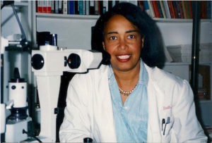 Dr. Patricia bath wearing a lab coat and sitting beside an eye examination instrument.