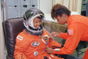 Two African-American women. One woman, Mae Jemison, is sitting wearing a space suit and helmet, smiling at the camera. The second woman, Sharon McDougle, is helping prepare Mae's space suit.