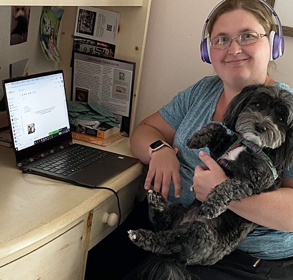 A woman wearing purple headphones holding a small gray dog on her lap. She is smiling at the camera and sitting beside an open laptop.